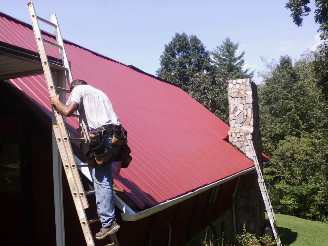 Roofer on a ladder beside a red metal roof and stone chimney — Soffit & Fascia Repair by Roofing Brothers in Hazel Crest